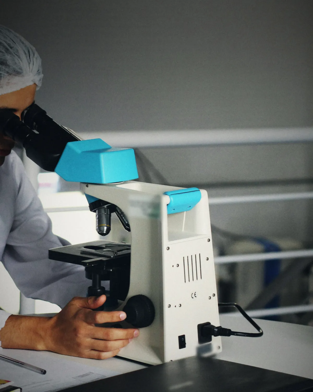 A lab worker in a hairnet looks through a microscope