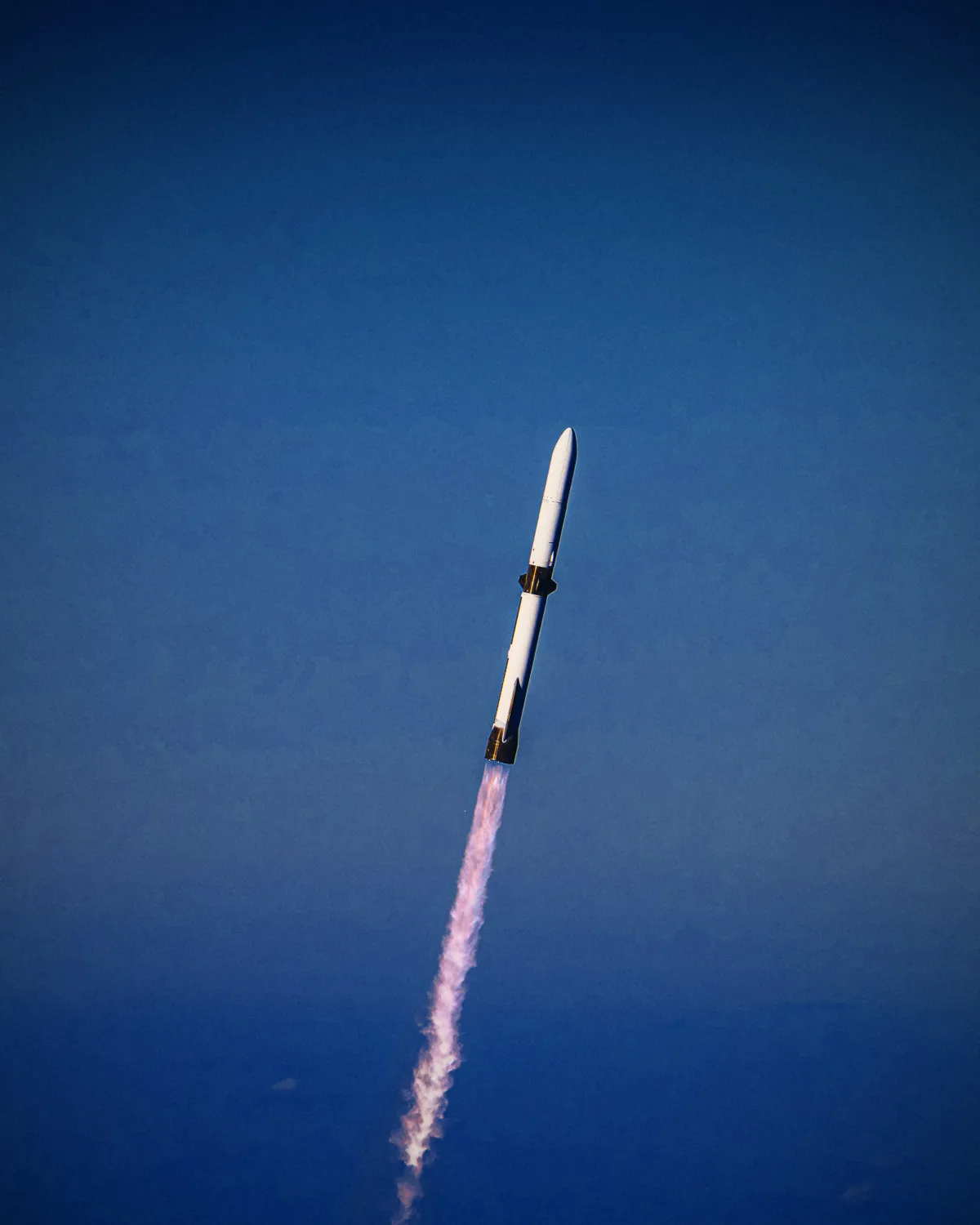 A white rocket climbs through a deep blue sky on a trail of exhaust